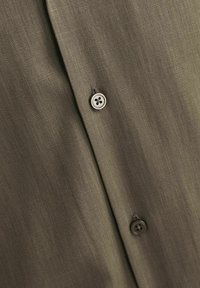 Tamsiai alyvuogių žalia marškiniai, pasižymintys minkšta, tekstūrine medžiaga su subtiliomis juostelėmis ir dviem apvaliais mygtukais priekyje, akcentuojančiais jų dizaino ypatybes.