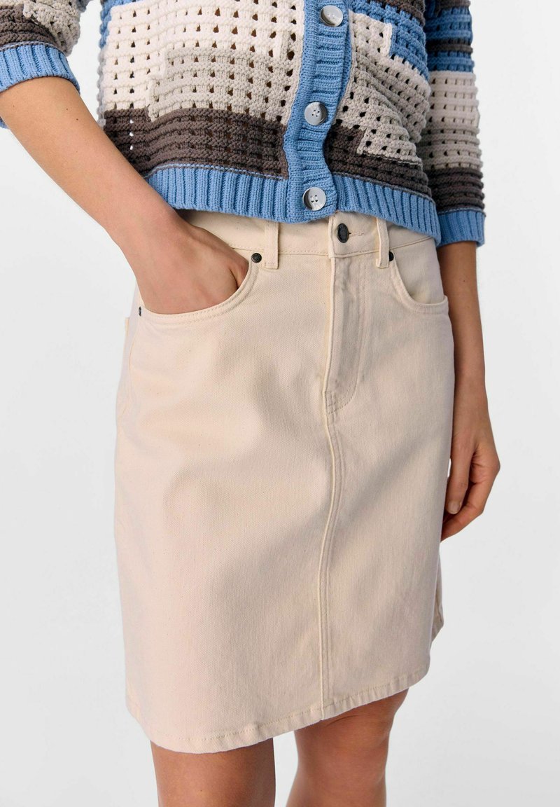 Beige denimrok met een rechte snit, voorzien van voorzakken en een zwarte knoopsluiting. De stof heeft een gladde textuur.