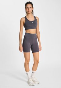 Grijze sportieve crop top en hooggereree shorts, beide van een gladde, rekbare stof, met een logo op de top. Draagt witte sokken en sneakers.