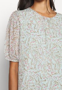 Jasnoniebieska bluzka w kwiaty z bufkami, zdobiona delikatnym wzorem różowych kwiatów i brązowych liści na teksturowanej tkaninie.