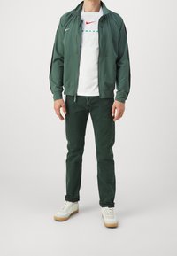 Vihreä vetoketjullinen takki mustilla yksityiskohdilla, valkoinen pyöreäkauluksinen t-paita, jossa on logo, vihreät teksturoidut housut ja valkoiset tennarit, joissa on kumipohja.