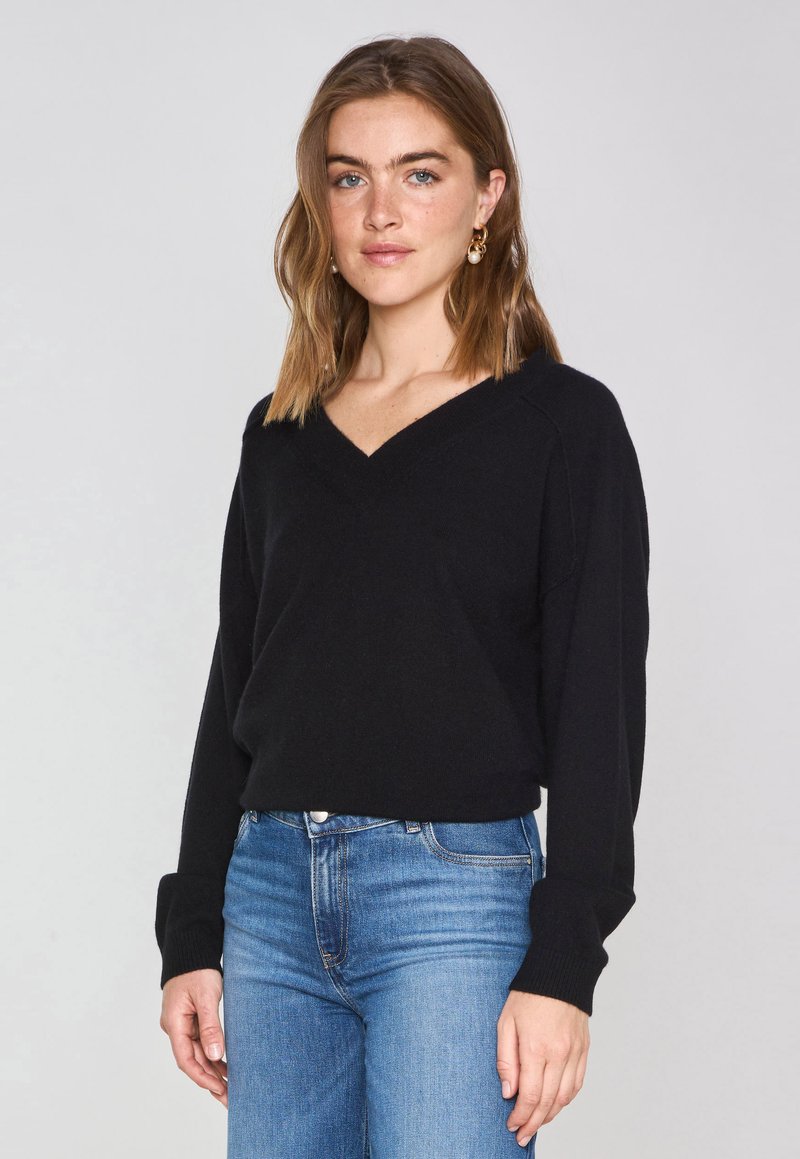 Pull noir à col en V avec manches longues, en matériau doux, porté avec un jean bleu ajusté, offrant une coupe décontractée et des poignets côtelés.