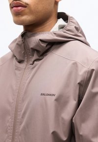 Κοντινό πλάνο ενός ατόμου που φοράει μωβ ζακέτα με φερμουάρ Salomon, με ανασηκωμένο γιακά και κουκούλα, μπροστά σε λευκό φόντο.