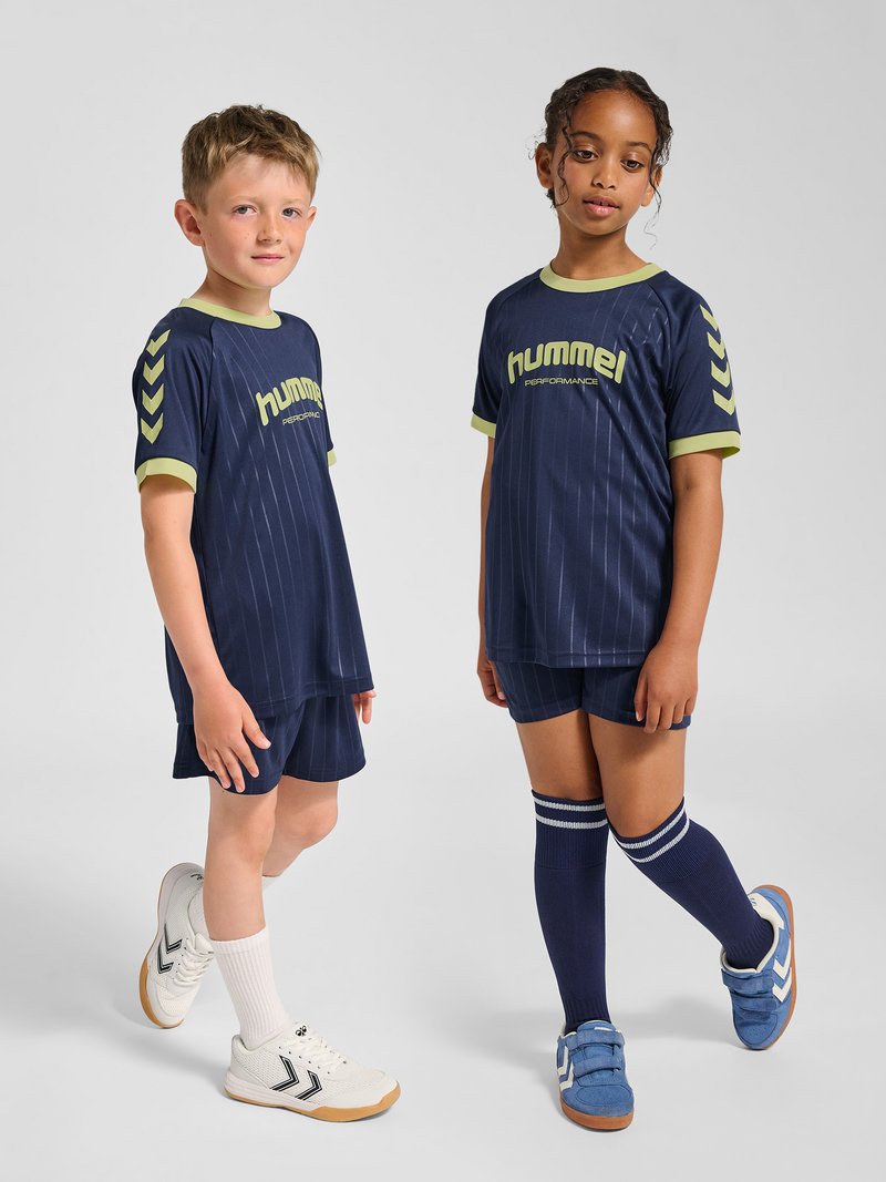 Twee kinderen dragen marineblauwe hummel sportkleding met korte broek en sneakers, staand op een effen lichtgrijze achtergrond.