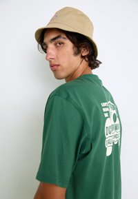 Grünes Baumwoll-T-Shirt mit kurzen Ärmeln, auf dem Rücken mit einem weißen grafischen Design versehen. Beigefarbener Bucket Hat, schräg getragen.