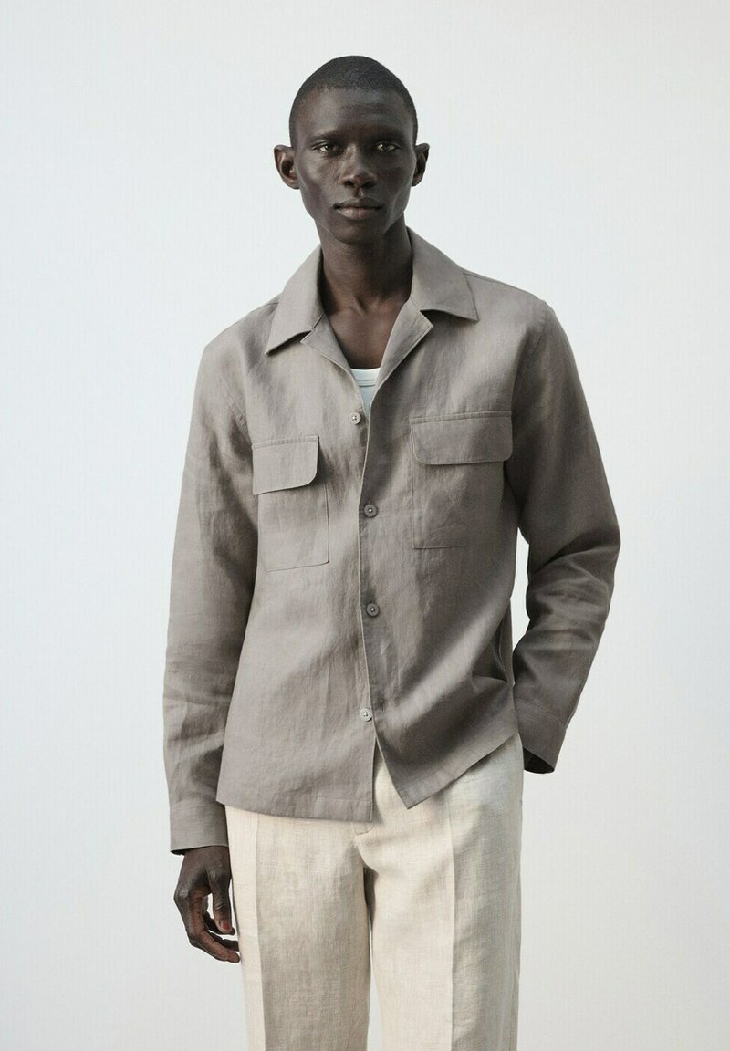 Homme portant une chemise à boutons gris clair sur un t-shirt blanc et un pantalon beige, debout contre un fond clair uni, une main dans la poche.