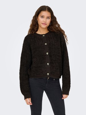 Kvinde med krøllet hår iført en sort tekstureret cardigan med knapper og mørke jeans, stående foran en ensfarvet lys baggrund.