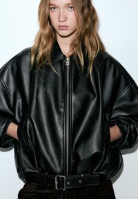 Fekete bőr bomberdzseki magas gallérral, túlságosan bő szabással és cipzáros elülső résszel. Derékrésznél övvel, ujjai struktúrált kialakításúak.