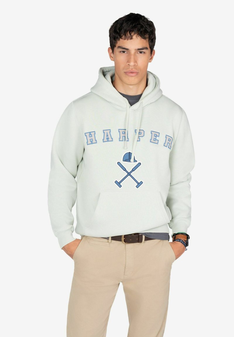 Sudadera verde claro con bolsillo frontal, que presenta "HARPER" en letras azules y un diseño de murciélagos cruzados. Lleva puesta con pantalones beiges y una camiseta en capas.