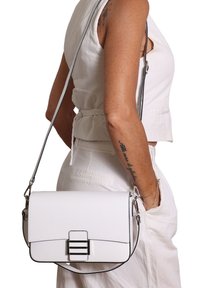 Donna che indossa un top senza maniche bianco e pantaloni, mentre porta una borsa a tracolla bianca con una fibbia metallica rettangolare.