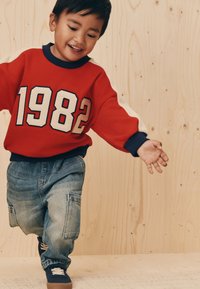 Sweat-shirt rouge avec "1982" en blanc, accents bleus, et jean ample. L'enfant porte des chaussures bleu marine avec des lacets blancs.