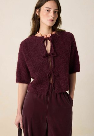 Burgunderfarbener Strickcardigan mit kurzen Ärmeln, ausgestattet mit einem Schlüsselring-Ausschnitt und kontrastierenden Bändern. Kombiniert mit passenden burgunderfarbenen Satin-Hosen.