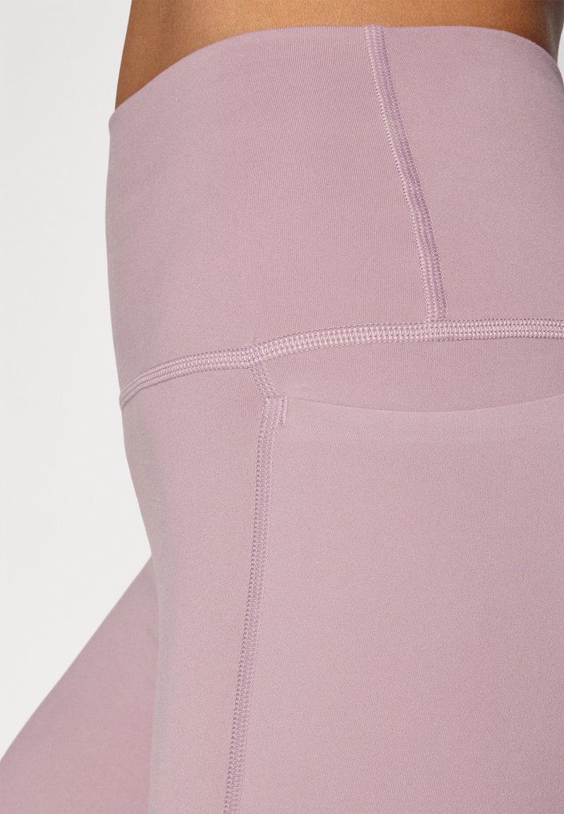 Leggings taille haute en tissu lilas doux, présentant une texture lisse et des coutures contrastées le long des seams, avec un détail de poche sur le côté.