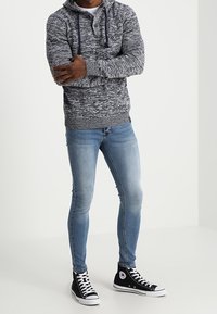 Felpa grigia a maglia con cappuccio e coulisse, abbinata a jeans skinny blu e sneaker alte nere con dettagli bianchi.