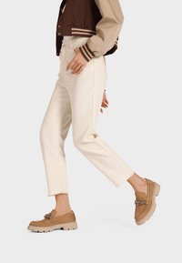 Mocassini beige in suede con suola chunky, caratterizzati da hardware decorativo e punta arrotondata. Indossati con jeans bianchi a vita corta e una giacca marrone.