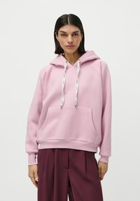 Femme aux cheveux raides et foncés portant un hoodie rose avec des cordons blancs et un pantalon large rouge foncé, se tenant devant un fond blanc.