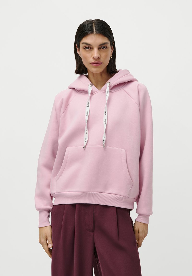 Donna con capelli lunghi e lisci scuri, che indossa un felpa rosa con cappuccio e stringhe bianche e pantaloni ampi rossi scuri, in piedi su uno sfondo bianco.