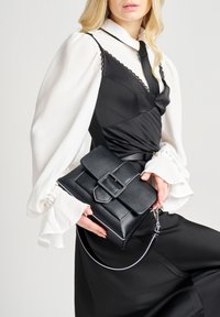 Borsa a mano in pelle nera con superficie texturizzata, forma rettangolare, dettaglio con fibbia e tracolla removibile. Esposta con una blusa di colore chiaro.