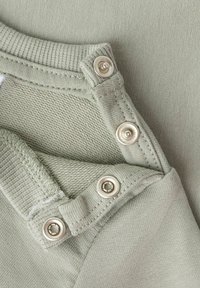 Zielona bawełniana tkanina z teksturowanym dzianym wzorem i gładkimi sekcjami. Widoczne metalowe zatrzaski z błyszczącym wykończeniem i wzmocnione szwy.