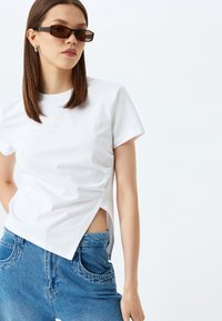 Camiseta blanca de manga corta con una abertura lateral, combinada con unos vaqueros de mezclilla azul de cintura alta. Modelo con gafas de sol rectangulares oscuras.
