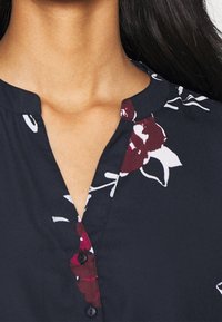 Marineblouse met een bloemenpatroon; heeft een ronde halslijn, knoopsluiting en korte mouwen. Stof lijkt glad met een zachte textuur.