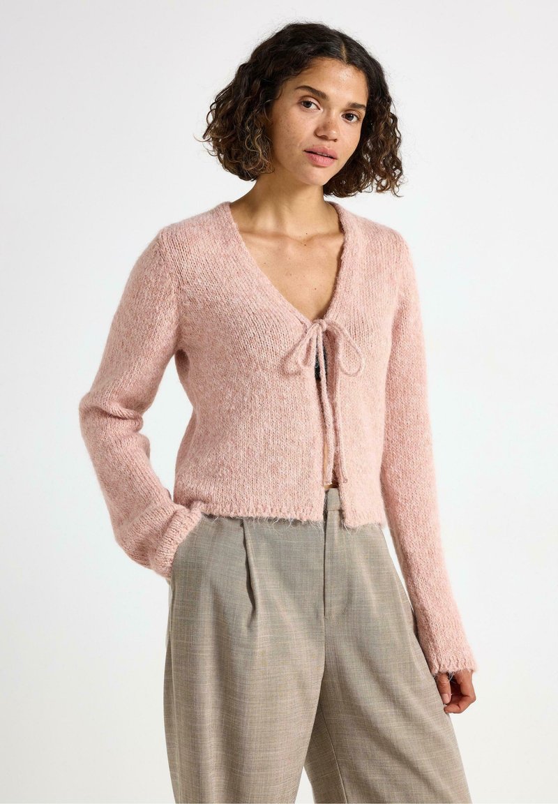 Kvinna med lockigt hår som bär en rosa stickad kofta knuten framtill och beige högmidjade vida byxor, står med ena handen i fickan.