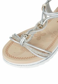 Sandalo in stile gladiatore argento con cinture decorate, fondo beige testurizzato e suola bianca glitterata. Presenta dettagli metallici rotondi.