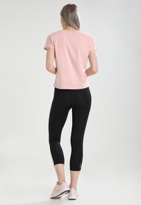 Žena s dlouhými blond vlasy, oblečená v růžovém tričku, černých krátkých legínách a růžových teniskách, stojící zády k fotoaparátu na jednobarevném pozadí.