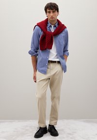 Chemise boutonnée à rayures bleu clair et blanches, pantalon crème, chaussures noires, et un pull en laine rouge drapé sur les épaules.
