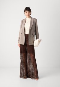 Pārbaudīts blazeris ar dubultās pogas dizainu, krēms turtlenecks un brūni vaļīgas adītas wide-leg bikses. Modelis tur krēma krāsas somu.