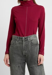 Bordeaux lange mouwen top met een hoge kraag en zilveren stiksels; gecombineerd met hoog getailleerde grijze denim broek met patchzakken.