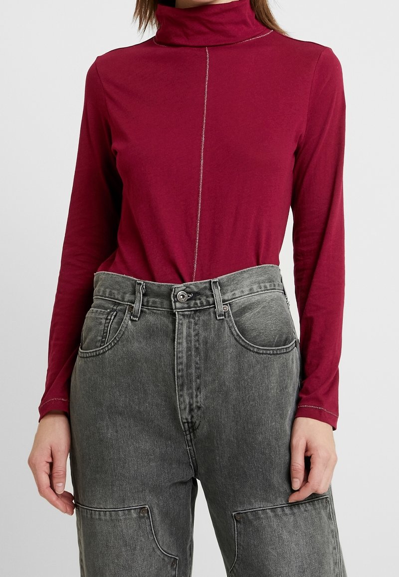 Bordeaux lange mouwen top met een hoge kraag en zilveren stiksels; gecombineerd met hoog getailleerde grijze denim broek met patchzakken.