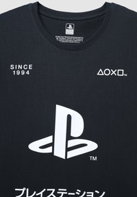 Maglietta nera in cotone con un grande logo PlayStation bianco, testo "SINCE 1994" e caratteri giapponesi. Scollo rotondo e etichetta visibile.