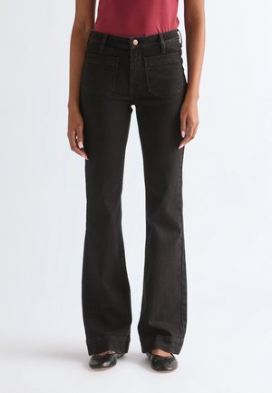 Persoon die hooggetailleerde zwarte flare jeans met frontzakken draagt, gecombineerd met zwarte ballerina's en een rood shirt, staande tegen een witte achtergrond.