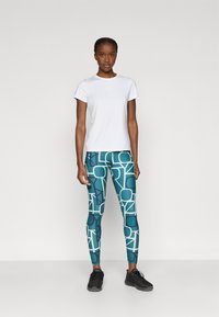 Λευκό αθλητικό πουκάμισο με κοντά μανίκια συνδυασμένο με τιρκουάζ κολάν που φέρουν γεωμετρικό μοτίβο σε ανοιχτό και σκούρο μπλε. Μαύρα αθλητικά παπούτσια.