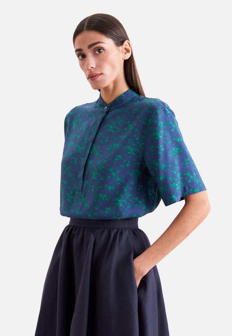 Femme aux cheveux raides et foncés, portant un chemisier bleu à manches courtes fleuri et une jupe haute foncée, une main dans la poche.
