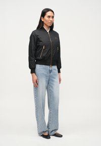 Μαύρο μπουφάν bomber με χρυσά φερμουάρ, φορεμένο πάνω από ανοιχτό μπλε τζιν παντελόνι με φαρδιές παντελόνες. Απλός σχεδιασμός με ριμπ τελείωμα και μανσέτες. Μινιμαλιστική εμφάνιση.