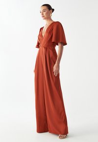 Mono de color óxido con escote en V profundo, mangas flotantes y piernas anchas. Tejido suave y pliegues que acentúan la cintura. Joyería con acentos dorados.