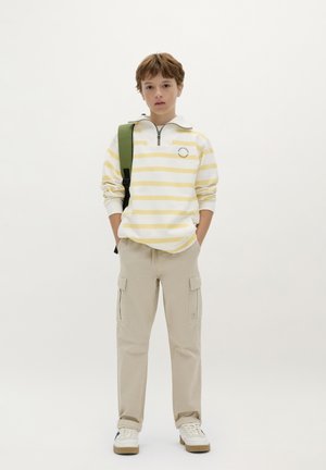Weiß und gelb gestreifter Sweatshirt mit Reißverschluss und Stehkragen, beige Cargo-Hosen und weißen Sneakers; grüner Rucksack über einer Schulter getragen.