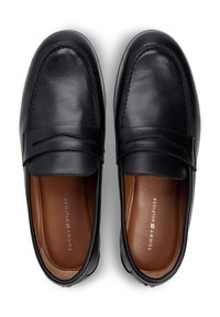 Μαύρα δερμάτινα μοκασίνια με λεία επιφάνεια, διακοσμημένο λουράκι στο πάνω μέρος. Καφετί δερμάτινη εσωτερική επένδυση με λεπτομέρεια λογότυπου στην ενδιάμεση σόλα.