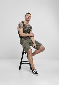 Maskáčová tílko, khaki cargo šortky s kapsami, sedící na černé stoličce, obuté černé tenisky, ukazuje tetování na paži.
