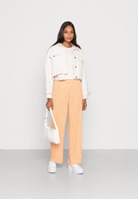 Jaqueta de denim curta branca sobre uma blusa rosa pastel, combinada com calças largas amarelas com padrão floral e ténis brancos de plataforma. Está a segurar uma bolsa branca.