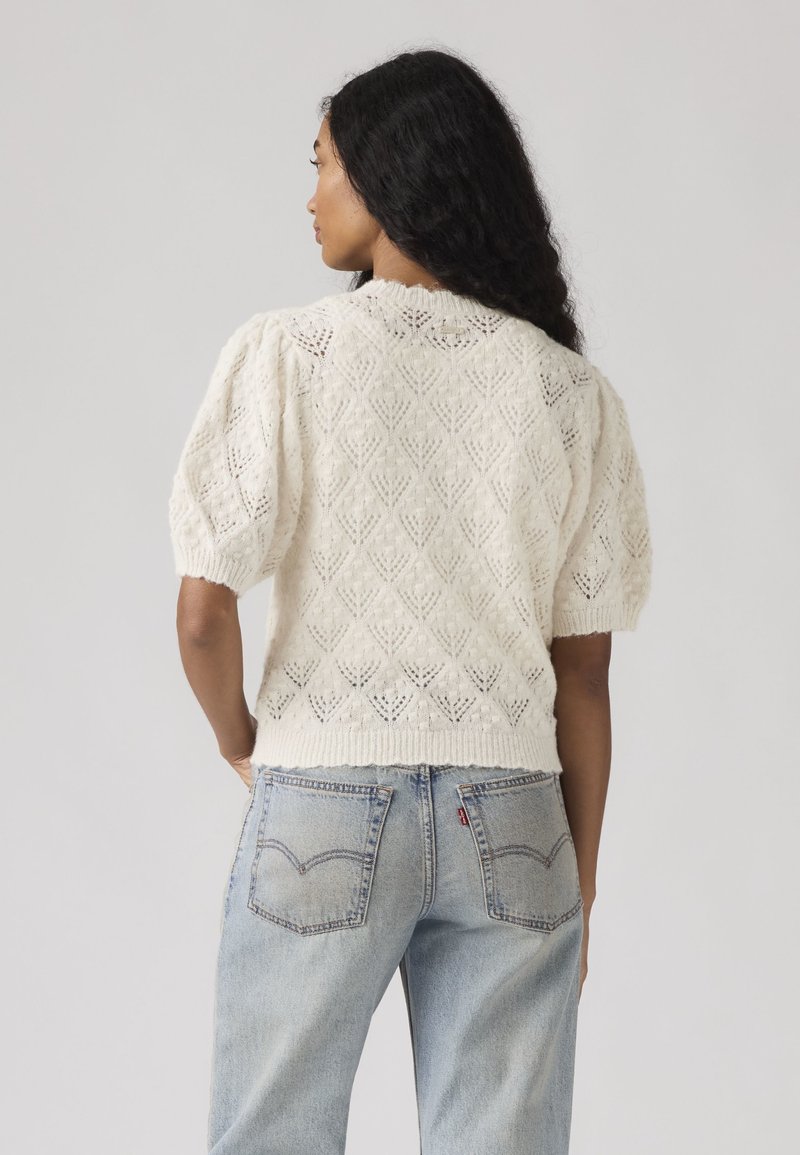 Woman with long curly hair wears a white short-sleeve knit sweater with a lace pattern and light blue denim jeans, facing away.