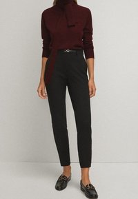 Top bourgogne à manches longues avec un col noué, associé à un pantalon noir ajusté et des mocassins noirs brillants avec des accents en hardware doré.