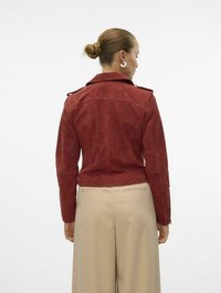 Rode suède jas met een kraag en schouderepauletten, gecombineerd met beige wijde broek. Gladde textuur, getailleerd ontwerp en geen zichtbare sluitingen.