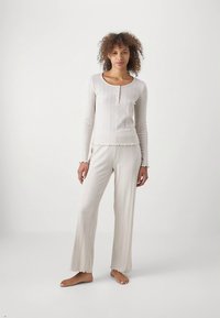 Šviesios smėlio spalvos poilsio komplektas su ilgomis rankovėmis, apvalios iškirptės ir sagų eilių palaidine, derinamas su plačiomis kelnėmis, abi su raukiniais krašteliais.