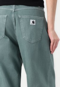 I pantaloni in denim verdi presentano due tasche posteriori con un'etichetta rettangolare, cuciture angolate e un fit rilassato, mostrando una superficie liscia.