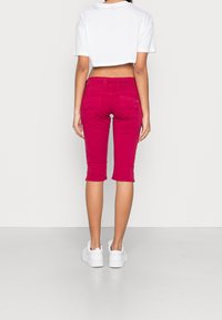 Camiseta blanca ajustada y recortada con pantalones rosa de largo hasta la rodilla. Los pantalones cuentan con dos bolsillos traseros con costuras onduladas. Se llevan con zapatillas blancas.