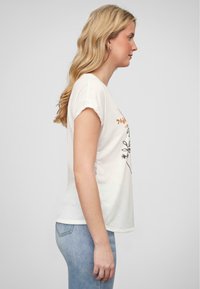 T-shirt en coton blanc avec manches retroussées, présentant un motif floral noir et du texte doré sur le devant, associé à un jean bleu clair.