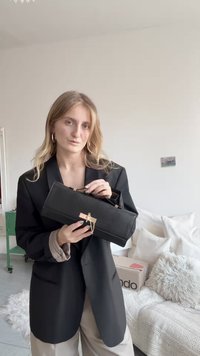 Fekete, téglalap alakú kézitáska texturált felülettel, arany színű fémrátétekkel és láncos díszítéssel. A modell fekete blézert és világos színű nadrágot visel.